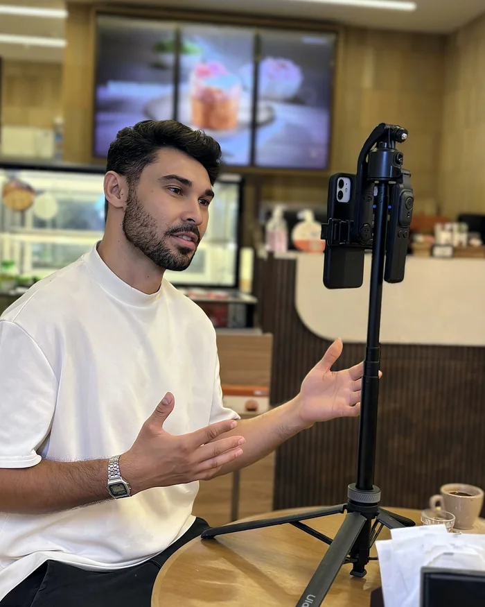 Homem sentado à mesa de um café, explicando algo com as mãos para o celular no tripé