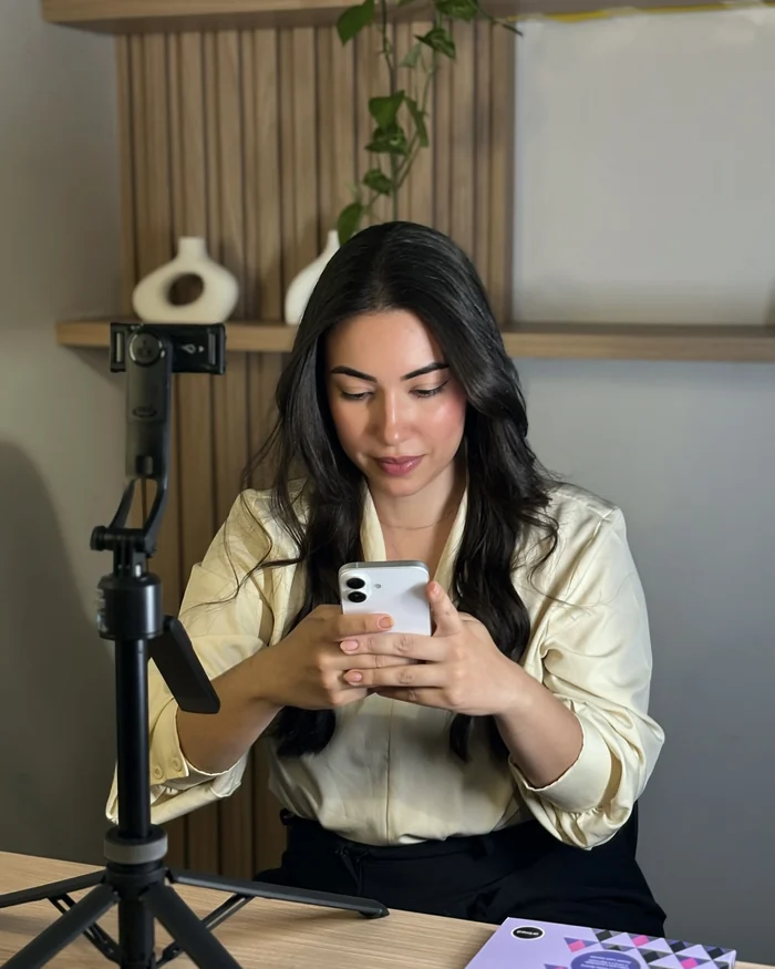 Mulher sentada à mesa, olhando o celular nas mãos, com o tripé ao lado
